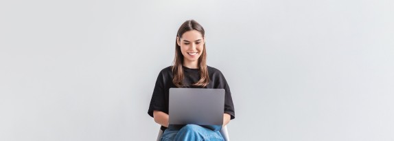 junge Frau sitzt auf einem Stuhl vor einer hellen Wand mit dem Laptop auf dem Schoß