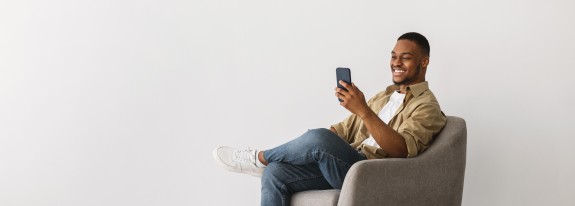 junger Mann sitzt auf einem Sessel vor einer hellen Wand mit dem Handy in der Hand